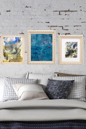 Three framed abstract paintings on a wall above a bed with pillows.