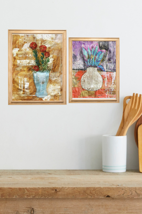 Two framed artworks on a wall above a wooden shelf with kitchen utensils.