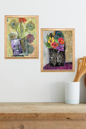 Two framed artworks of floral arrangements on a wooden shelf with a white wall background.