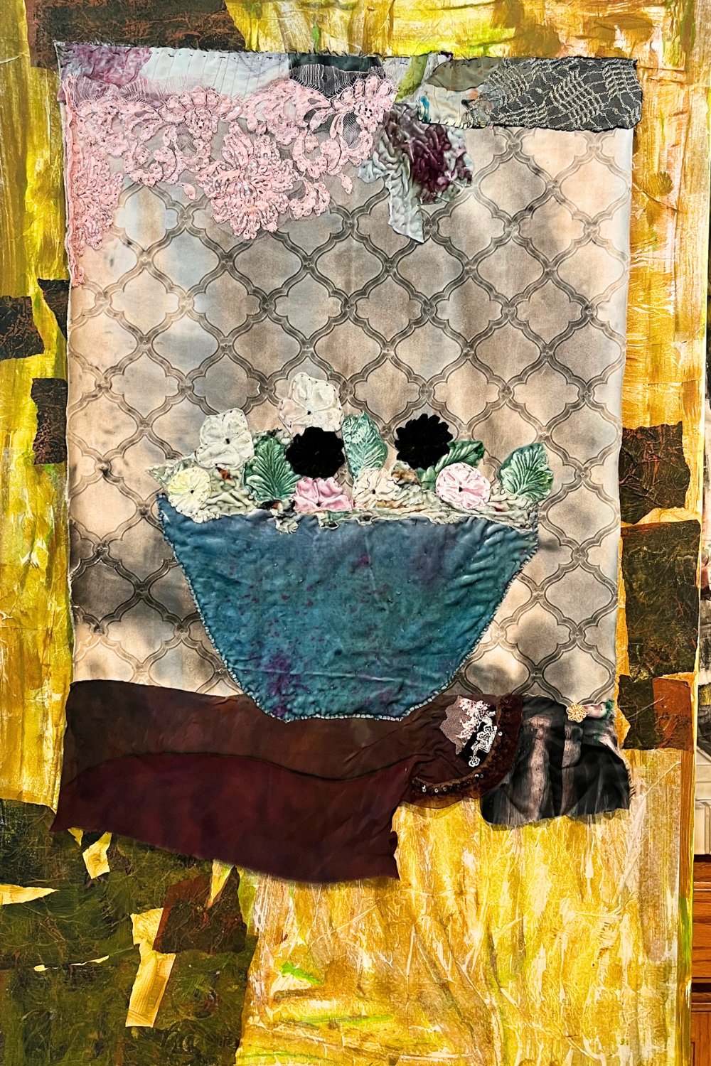 A vertical textile artwork or patchwork quilt featuring a stylized still life: a blue bowl with flowers, a beige patterned background, pink lace-like texture, dark brown and black fabric shapes, and a yellow-gold abstract border.