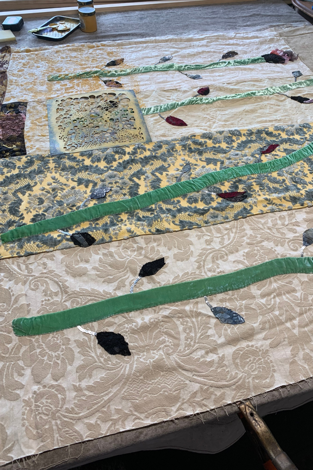 A close-up of a textile artwork or fabric panel laid flat on a table, featuring a beige damask background with layered green satin-like stripes, a yellow and gray patterned band, and scattered leaf-shaped fabric pieces in green, black, and red.