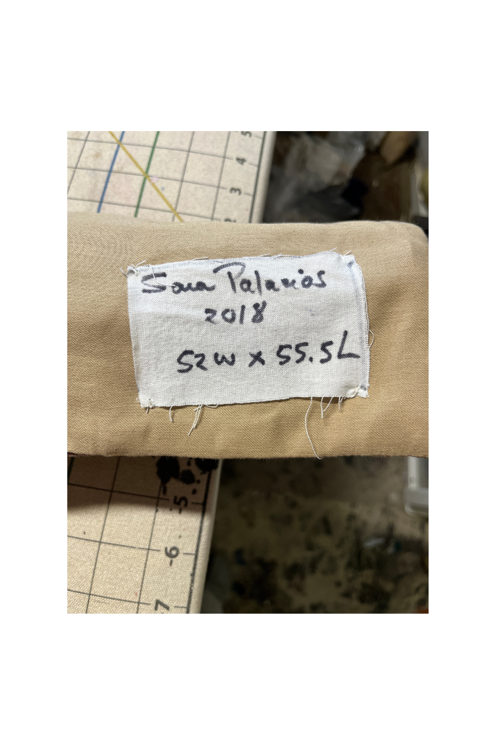 A close-up of a beige fabric item with a sewn-on white label reading 'Santa Palacios 2018 S2W x 55.5L' on a measuring board background.