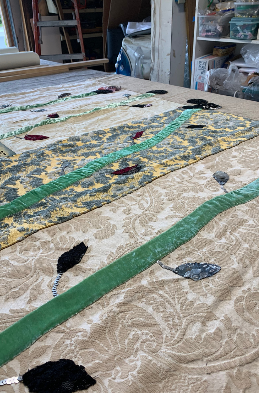 A large quilted textile piece laid out on a table in a workshop. The quilt features a beige damask background with green velvet border stripes, a yellow and gray patterned band, and scattered black and gray fabric patches.