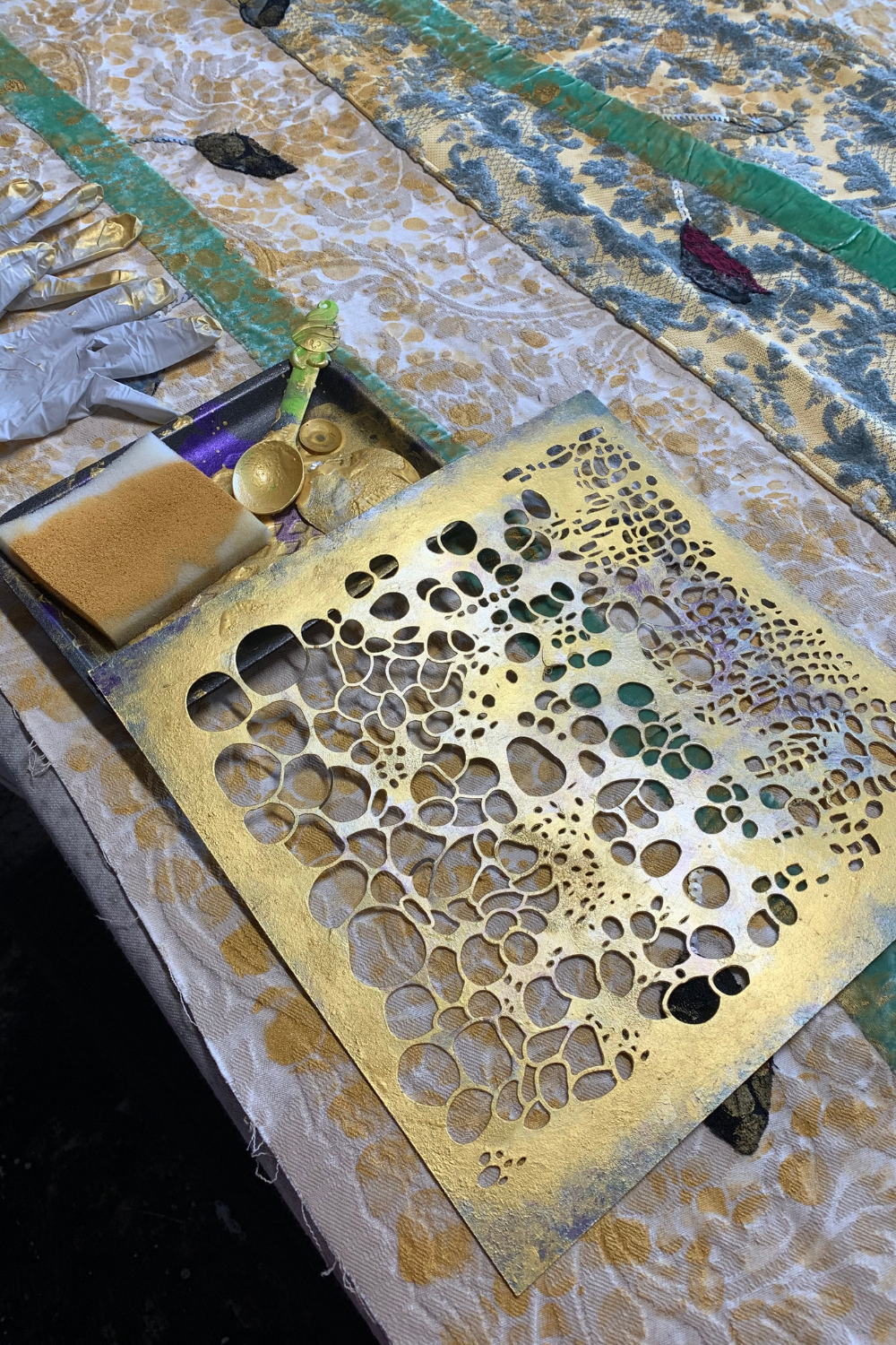 A square gold metal stencil with an organic bubble or coral-like cutout pattern, laid on a patterned craft table alongside paint, brushes, and other painting supplies.