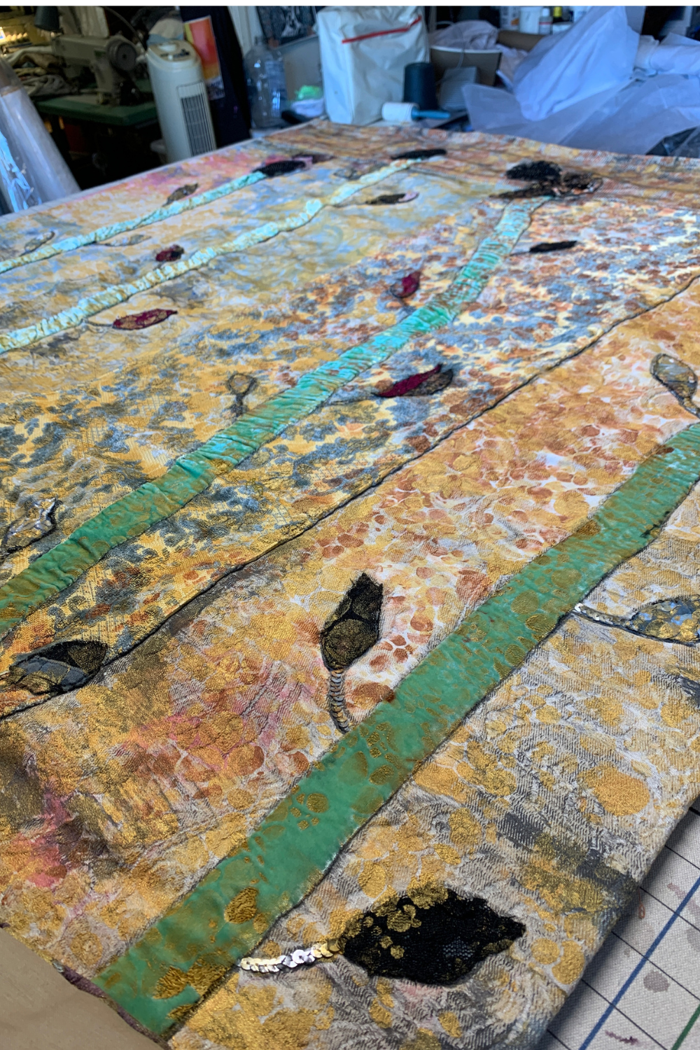 A large rectangular textile artwork or quilt laid flat on a table in a studio. The surface features an abstract mixed-media style design with gold, beige, brown, teal green, black, and pink areas, plus stitched leaf-like motifs and textured brushwork.