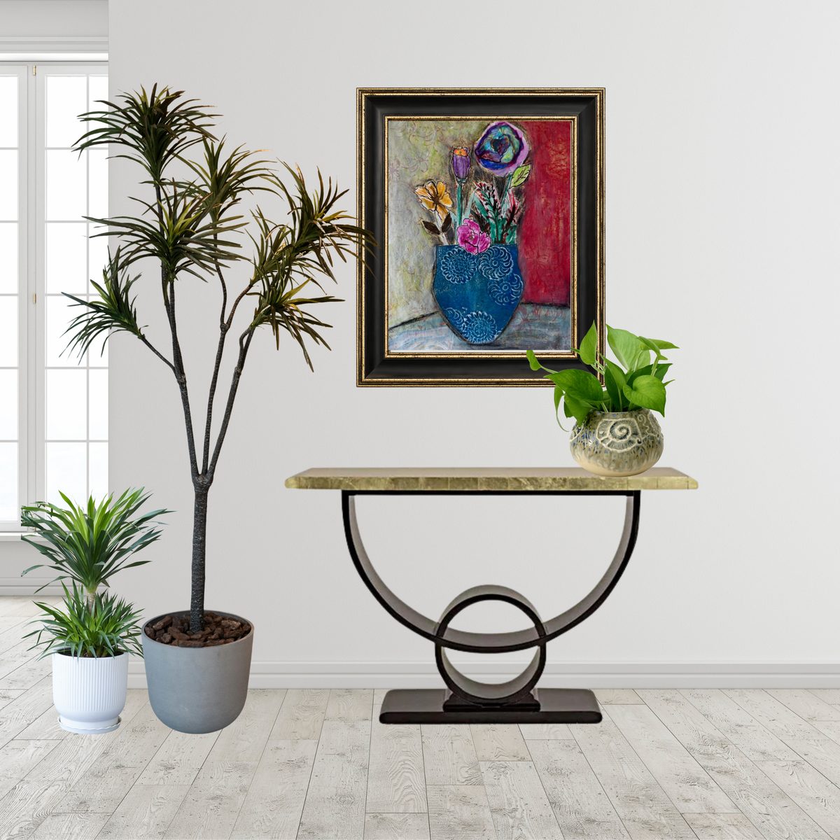 A framed still life painting of a blue vase with flowers hangs on a white wall above a console table with a stone top and curved metal base. Two potted plants are on the floor to the left, and a green plant sits on the table to the right.