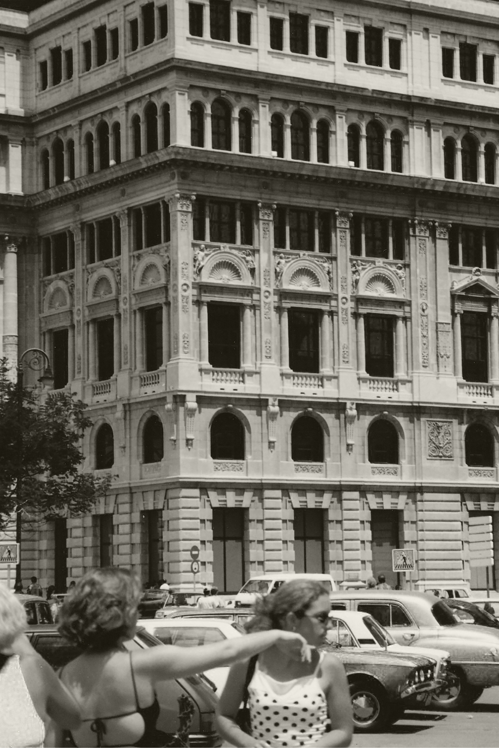 Two women standing in front of a large building with vintage cars in the foreground.
