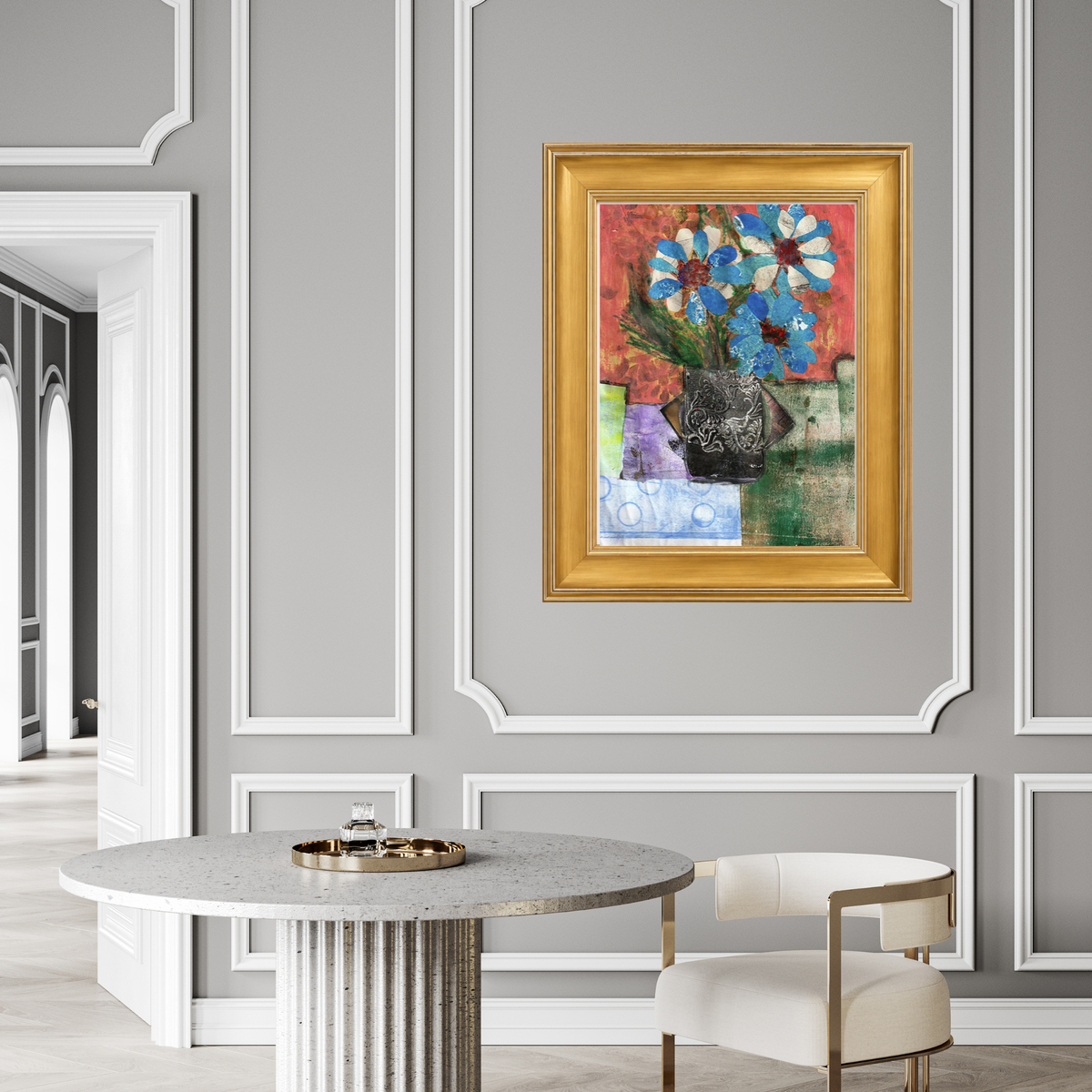 A framed abstract floral painting with blue, red, green, and purple brushstrokes inside a gold frame, displayed on a gray paneled wall above a round table and chair.