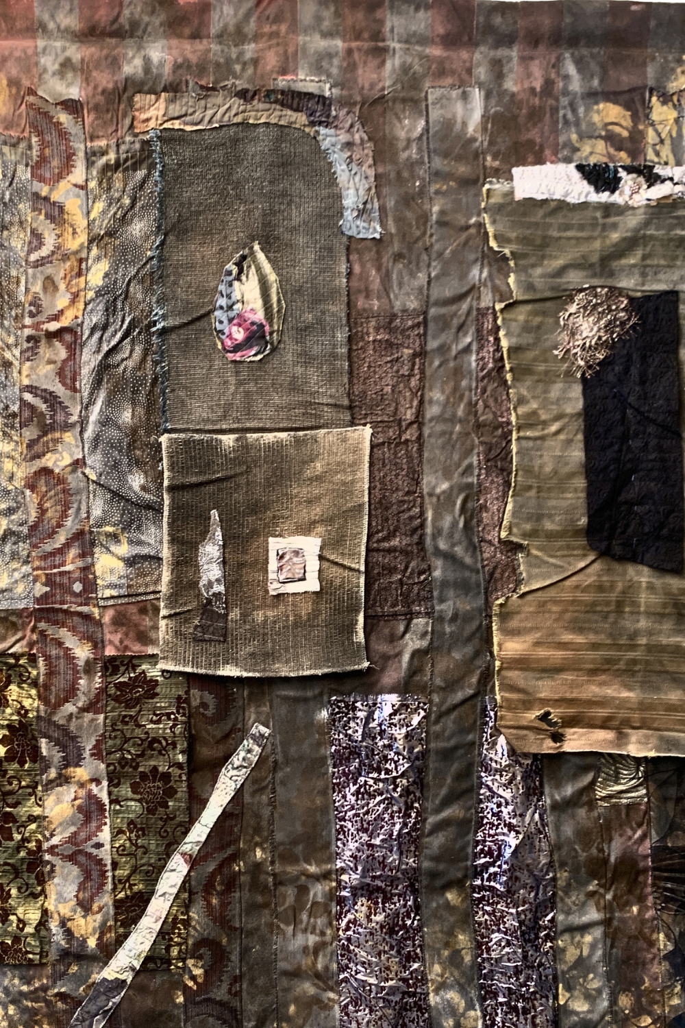 An abstract mixed-media textile artwork featuring layered patches of brown, beige, and black fabrics with distressed textures, torn sections, and a small central inset patch.