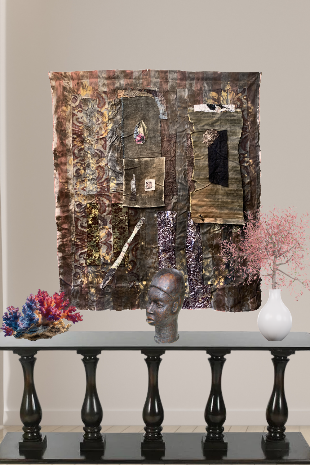 A large rectangular mixed-media wall art piece with layered paper, fabric, and textured panels in earthy brown, beige, and black tones, displayed above a black console table. The room setting includes a beige wall, a white vase with pink branches, a blue-and-pink coral-like object, and a sculptural head on the table.