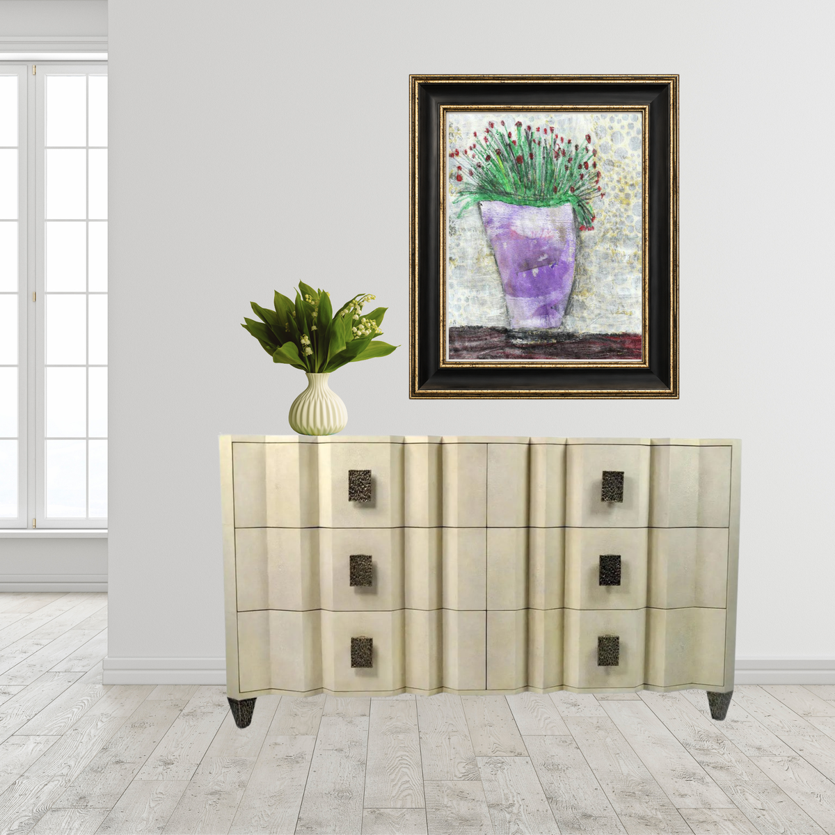 A framed art print depicting a purple vase with green plants, displayed on a light-colored wooden wall above a cream-colored credenza with dark metal knobs, next to a small potted plant.