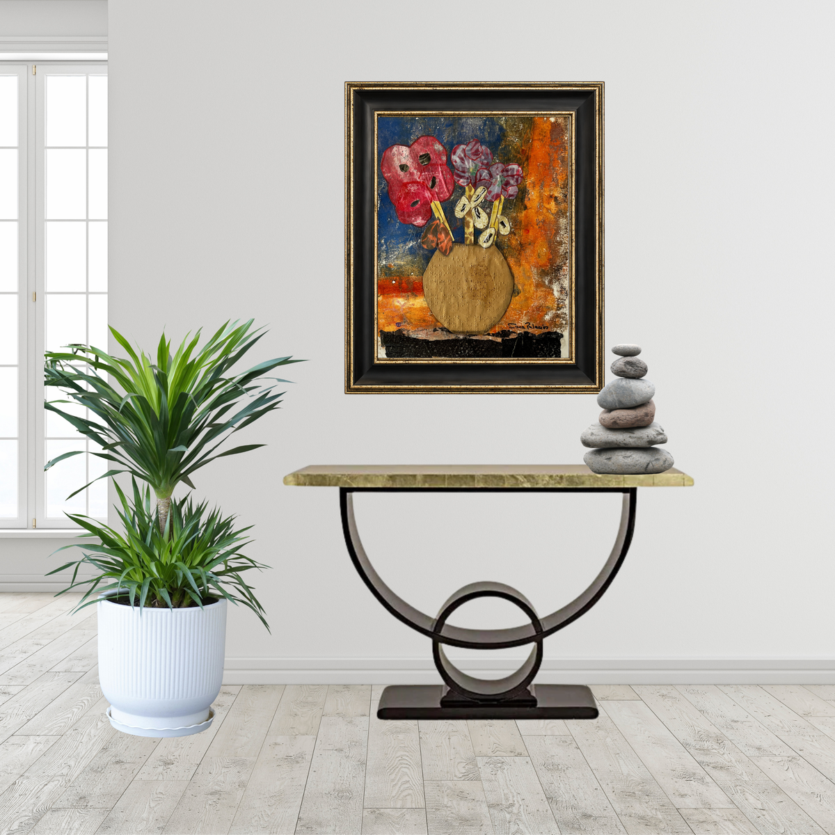 A framed abstract painting of flowers in a vase hangs on a white wall above a console table in a bright room with a potted plant and stacked stones.
